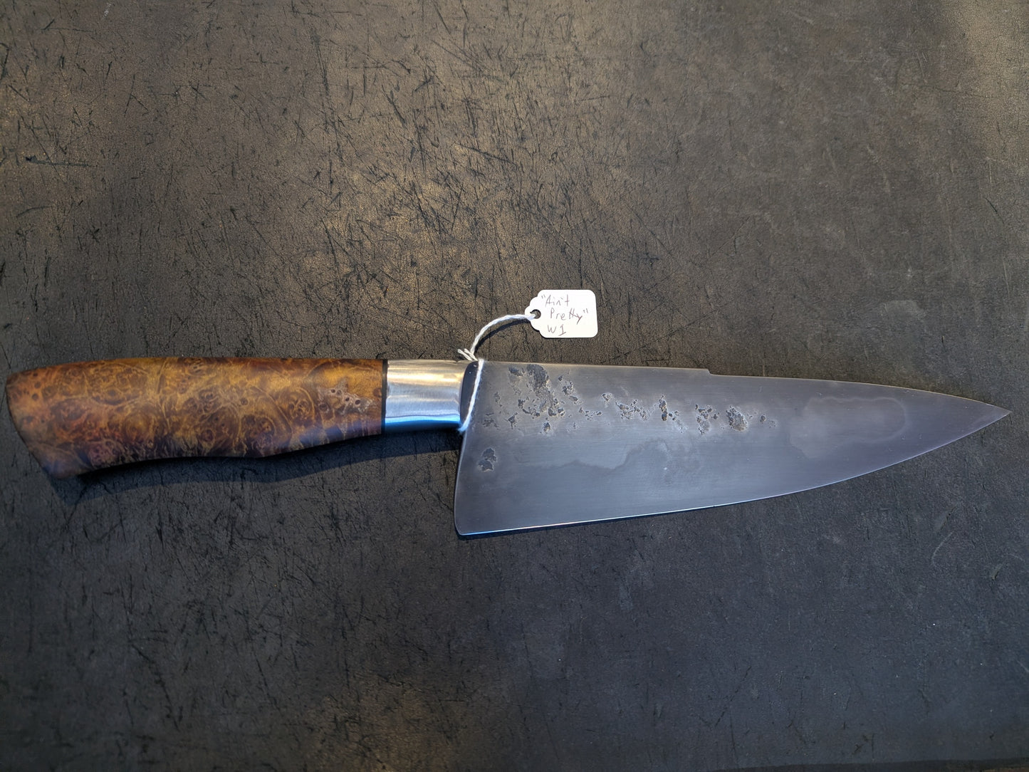 Black Cat Metal Mono Steel Chef knife with a burl wood handle, showcasing a patina and hammer marks, lies on a scratched surface with a Ain't Pretty tag.