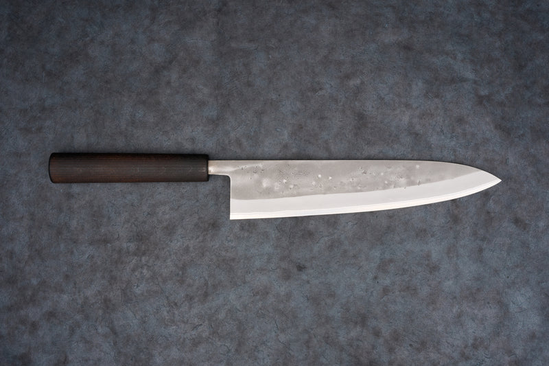 Fujimoto Nashiji Gyuto 240mm knife showcases a polished steel blade with hammer-textured pattern and dark wood handle, designed for precision slicing in Japanese cuisine, blending carbon and stainless steel attributes.