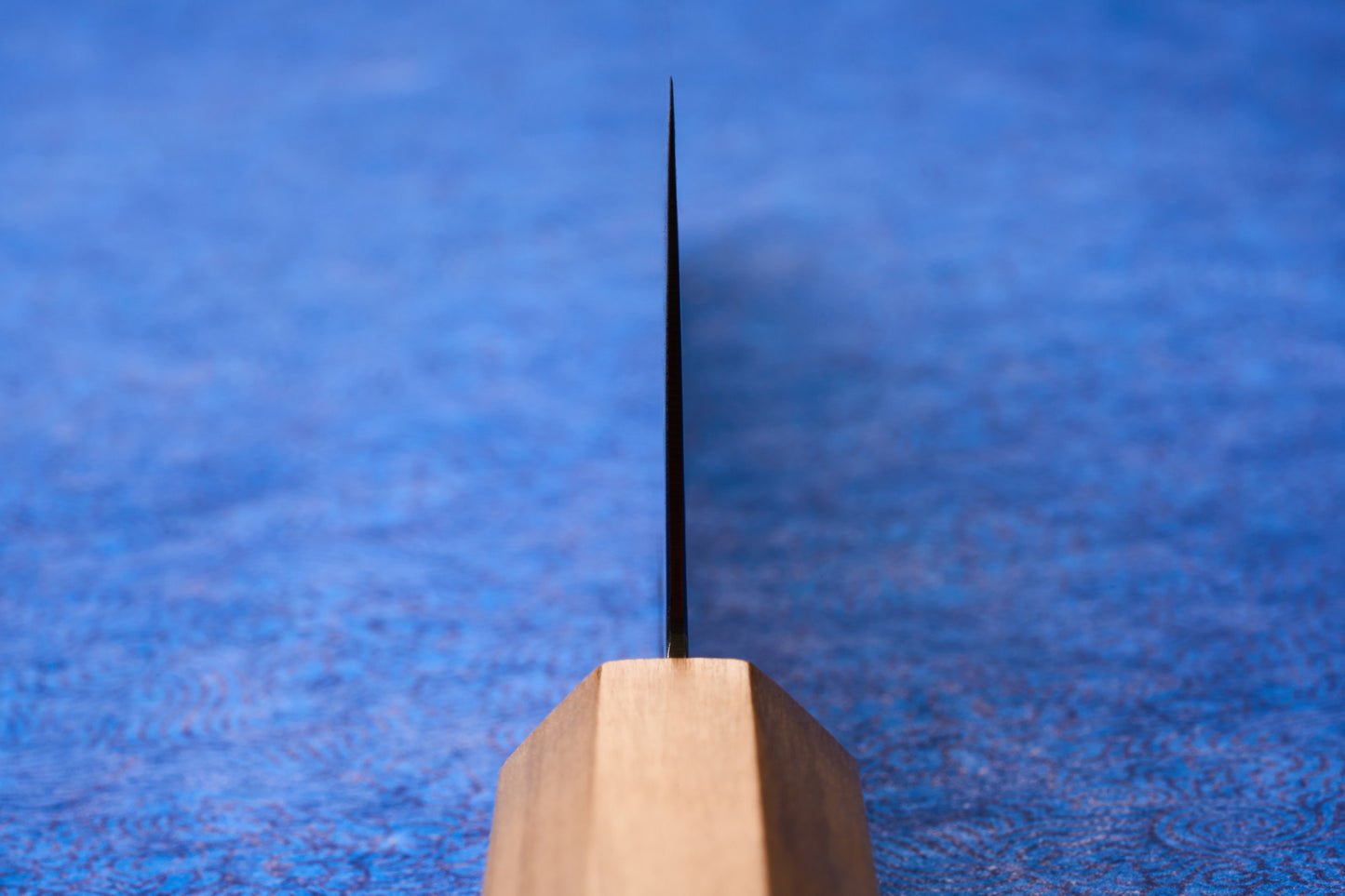 ICHIGO Haku Gyuto 240mm stands vertically, showcasing its thin metal blade and light wooden handle, set against a softly blurred blue background, embodying precision and elegance for culinary enthusiasts.