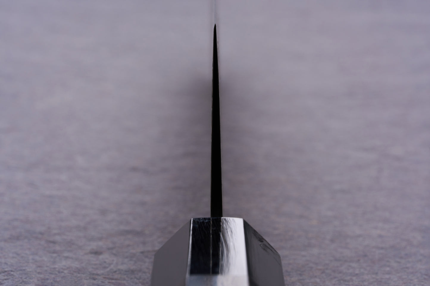 Close-up of Kanjo SG2 Nickel Damascus Kiritsuke Gyuto 240mm knife blade edge, showcasing its precision craftsmanship and sleek, polished bolster against a softly blurred tabletop backdrop.