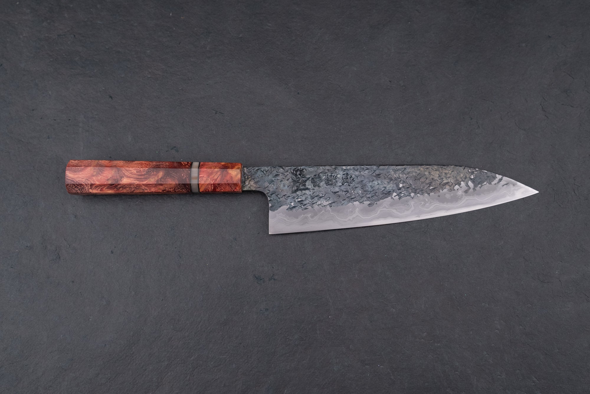Kisuke Manaka ENN Gyuto 210mm knife, featuring a hand-hammered Damascus-patterned blade and a richly figured wooden handle with a decorative spacer ring, showcased on a dark surface.