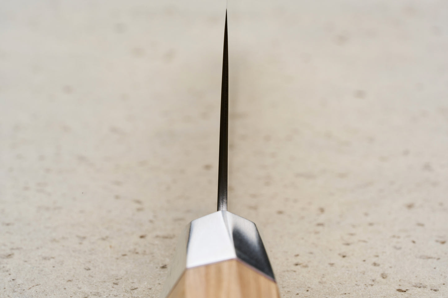 Mcusta Zanmai Beyond Kiritsuke Gyuto 210mm knife, showcasing its thin steel blade, metal bolster, and wooden handle, set edge-on against a light speckled surface, embodying precision craftsmanship.