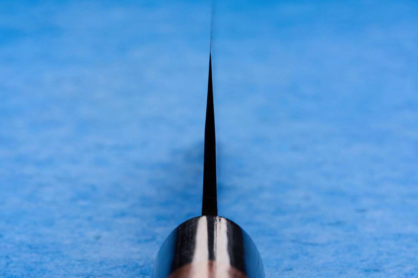Close-up of the Onihiro Shiroichi Kurouchi Nakiri 135mm, showcasing its sharp blade on a textured blue surface. The knife features a thin, dark blade tapering to a fine point, ideal for precise vegetable chopping.