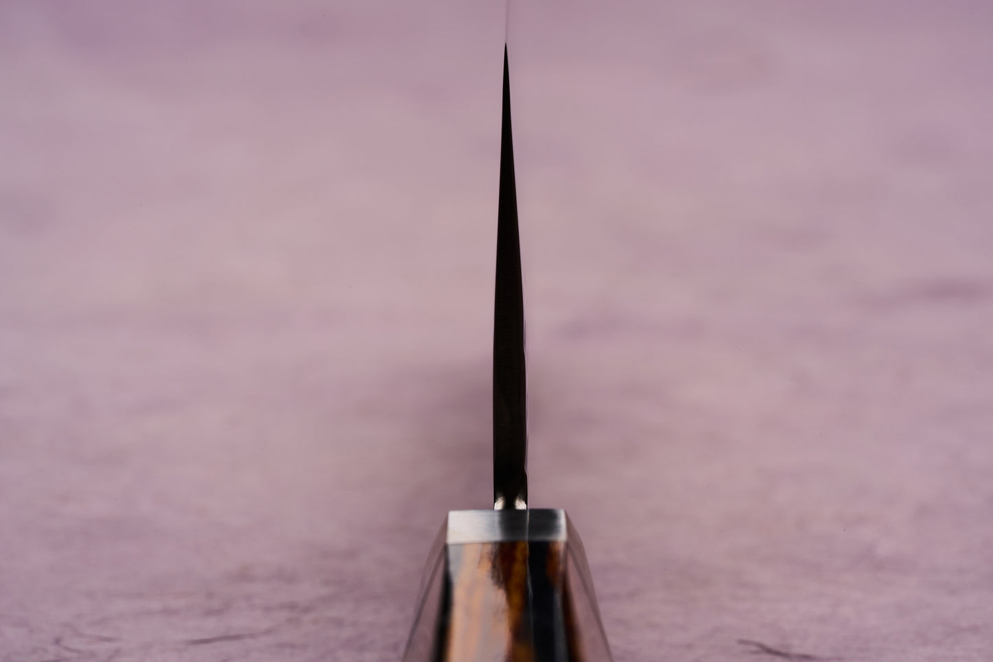 Ono Knife Suzukaze SG2 Tsuchime Damascus Santoku 180mm with a thin blade edge, wooden handle visible below, displayed against a soft pink background, showcasing precision craftsmanship and cutting-edge design.