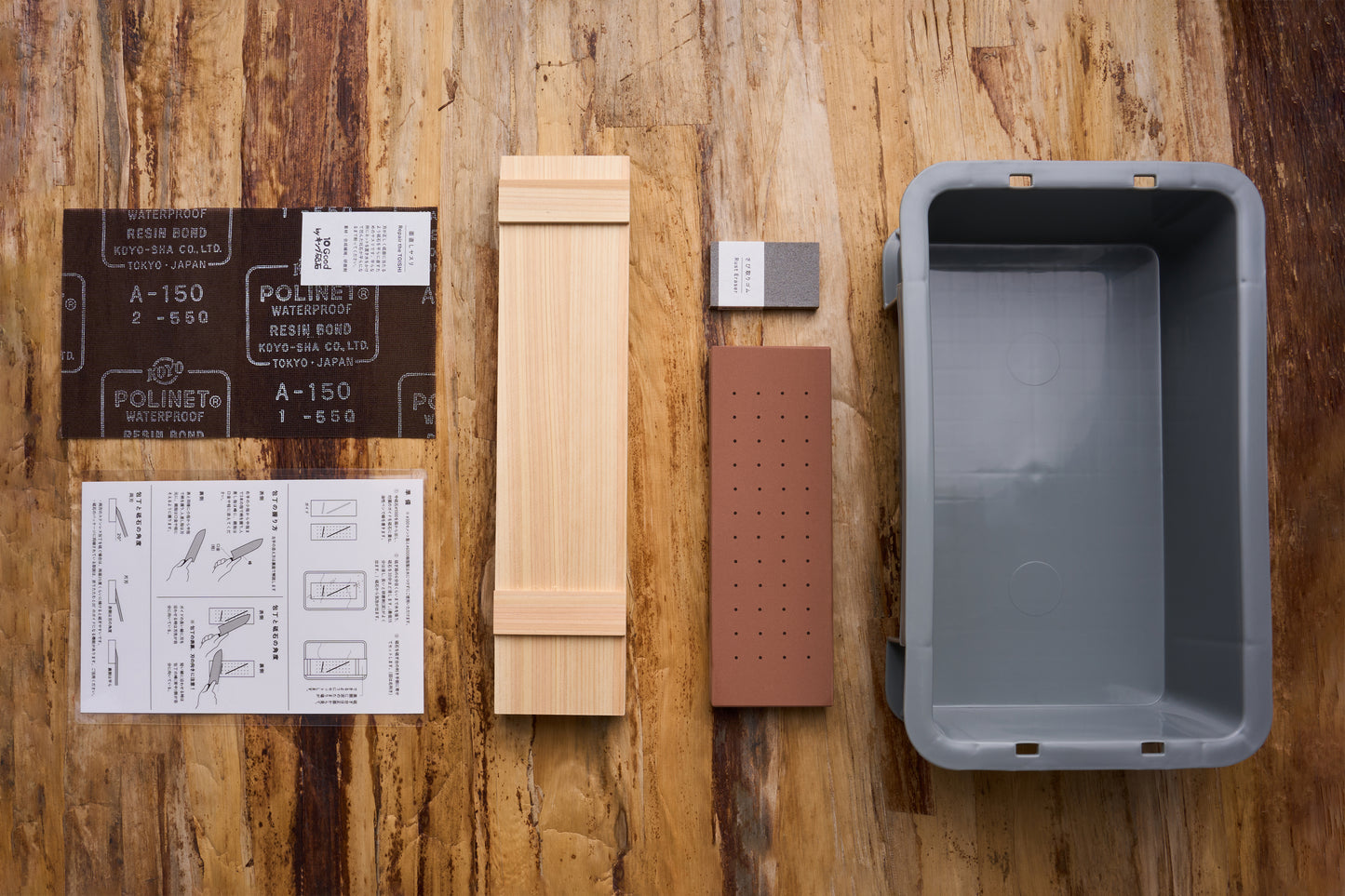 10 Good Togiba 5 Piece Set on table: abrasive paper, instruction sheet, wooden jig, grey stone, reddish-brown block, and basin for sharpening and maintaining knives.