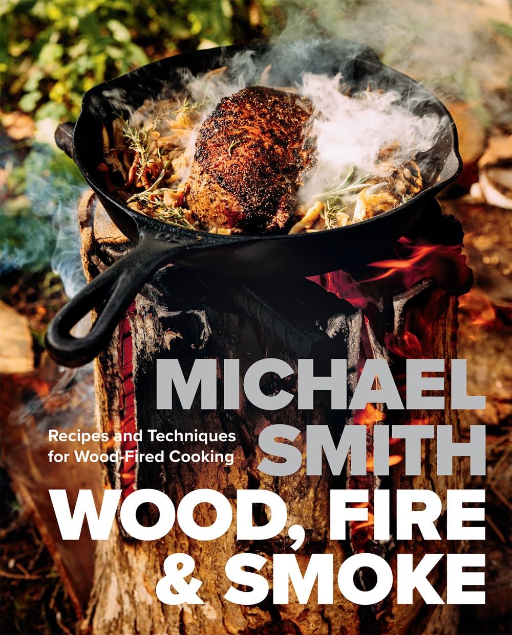 Cover of Wood, Fire & Smoke by Michael Smith, featuring a cast-iron skillet over a wood-fire with seared meat, herbs, and vegetables. Subhead: “Recipes and Techniques for Wood-Fired Cooking.” Ideal for culinary enthusiasts.