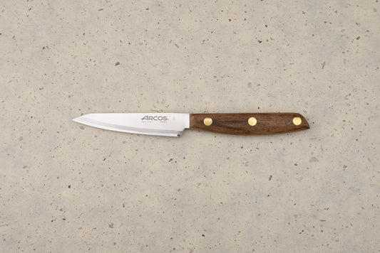 Arcos Nordika 100mm Petty Knife with a straight stainless-steel blade and dark wood handle, lying on a countertop. The blade features the Arcos logo and Made in Spain, Albacete engraving.