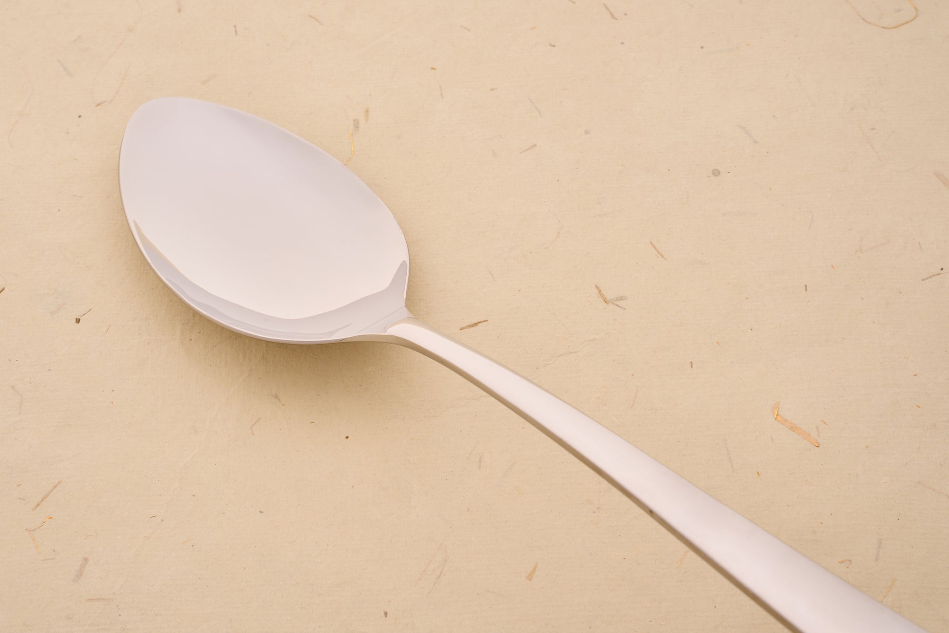 Arcos Berlin Series Serving Spoon close-up, showcasing its sleek metal design, reflecting the brand's centuries-old craftsmanship on a beige textured surface.