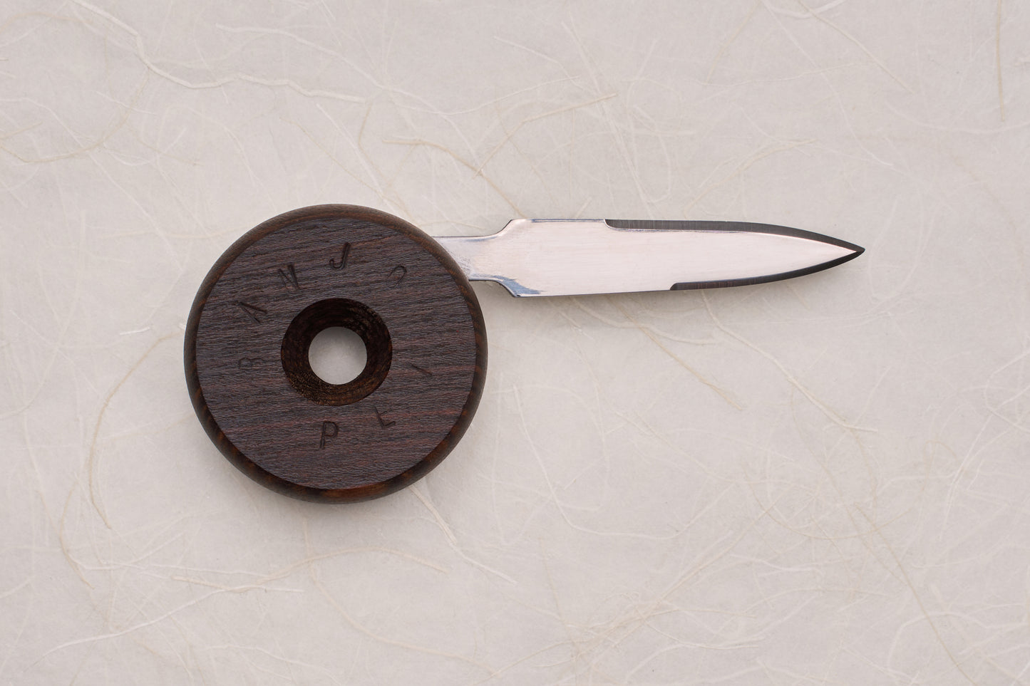 Banjo Oyster Knife with a circular dark wood handle and 2.5-inch precision stainless-steel blade, designed for ergonomic shucking. Handle features a finger hole and SAMPLE engraving, inspired by skateboard wheels.