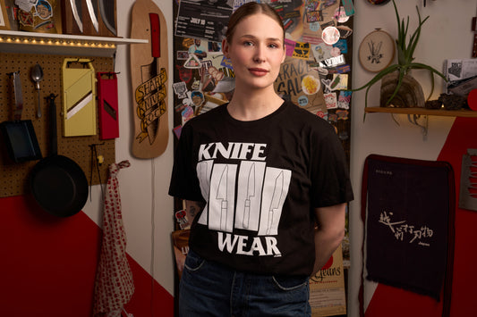 Knifewear Black Flag T-Shirt worn by a person in a workshop setting, featuring knife silhouettes and KNIFE WEAR print, surrounded by kitchen tools and culinary-themed decor.
