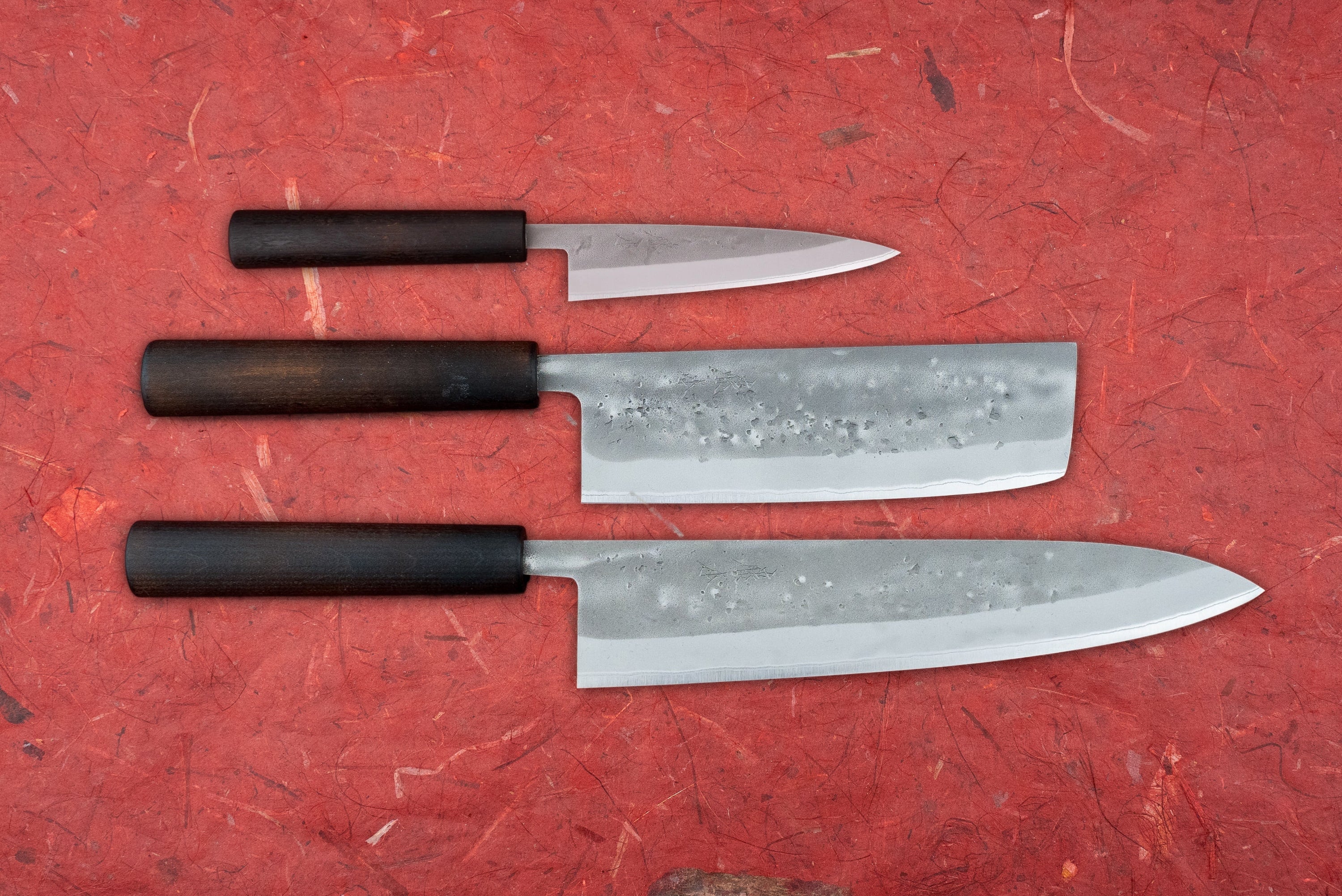 Fujimoto Nashiji 3 Piece Knife Set with Nakiri, showcasing a petty knife, nakiri, and gyuto, features hammered high-carbon steel blades and dark wooden wa handles on a red textured surface.