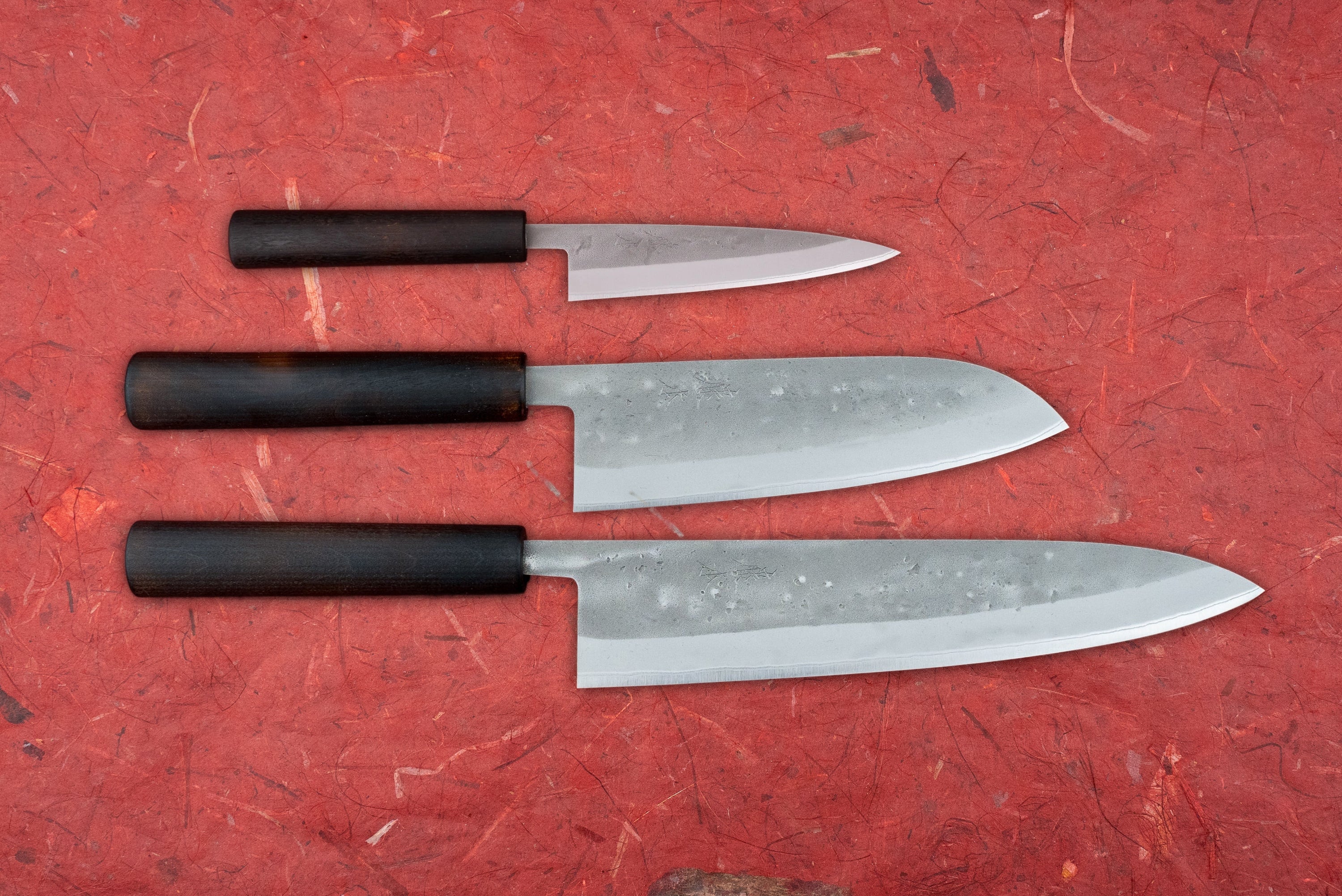 Fujimoto Nashiji 3 Piece Knife Set with Santoku, showcasing a paring, santoku, and gyuto knife, featuring hammered forged-steel blades and dark wooden handles, on a textured surface.