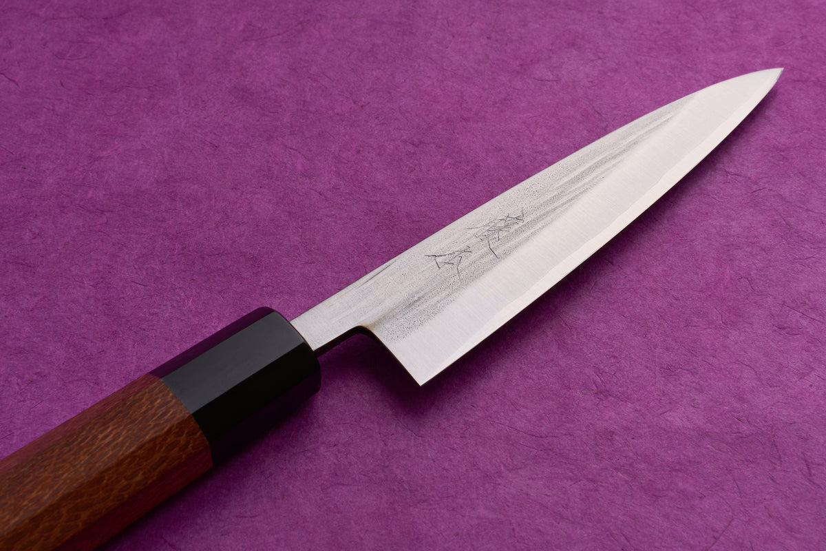 Fujimoto AS Sazanami Petty 135mm knife with a polished steel blade featuring engraved Japanese characters and an octagonal wooden handle, displayed on a textured purple surface.
