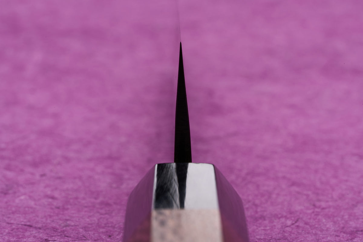 Fujimoto AS Sazanami Petty 135mm knife blade close-up, showcasing its sharp edge and polished spine, set against a textured pink background.