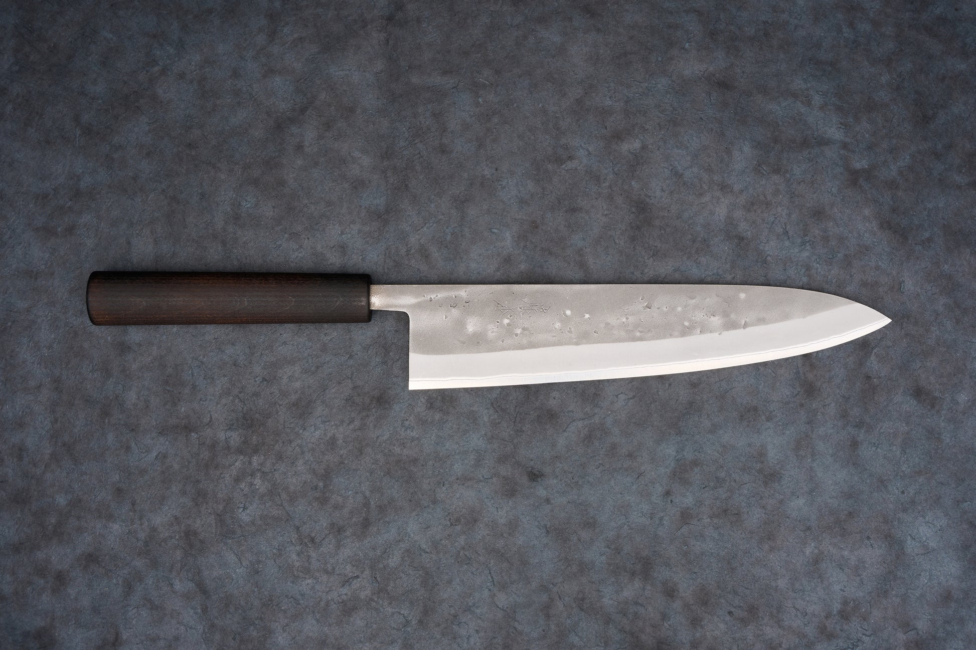 Fujimoto Nashiji Gyuto 240mm knife showcases a polished steel blade with hammer-textured pattern and dark wood handle, designed for precision slicing in Japanese cuisine, blending carbon and stainless steel attributes.