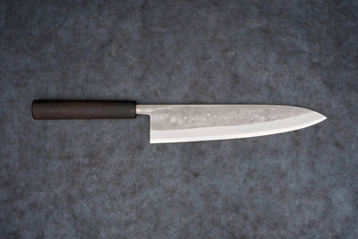 Fujimoto Nashiji Gyuto 240mm knife showcases a polished steel blade with hammer-textured pattern and dark wood handle, designed for precision slicing in Japanese cuisine, blending carbon and stainless steel attributes.