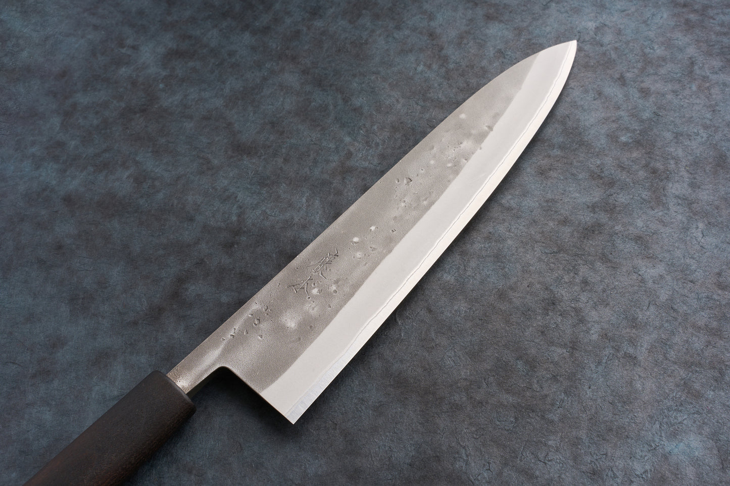 Fujimoto Nashiji Gyuto 240mm knife with a hammered finish, lying flat. The blade features a pronounced cutting edge and a visible dark handle, embodying the craftsmanship valued at Knifewear.