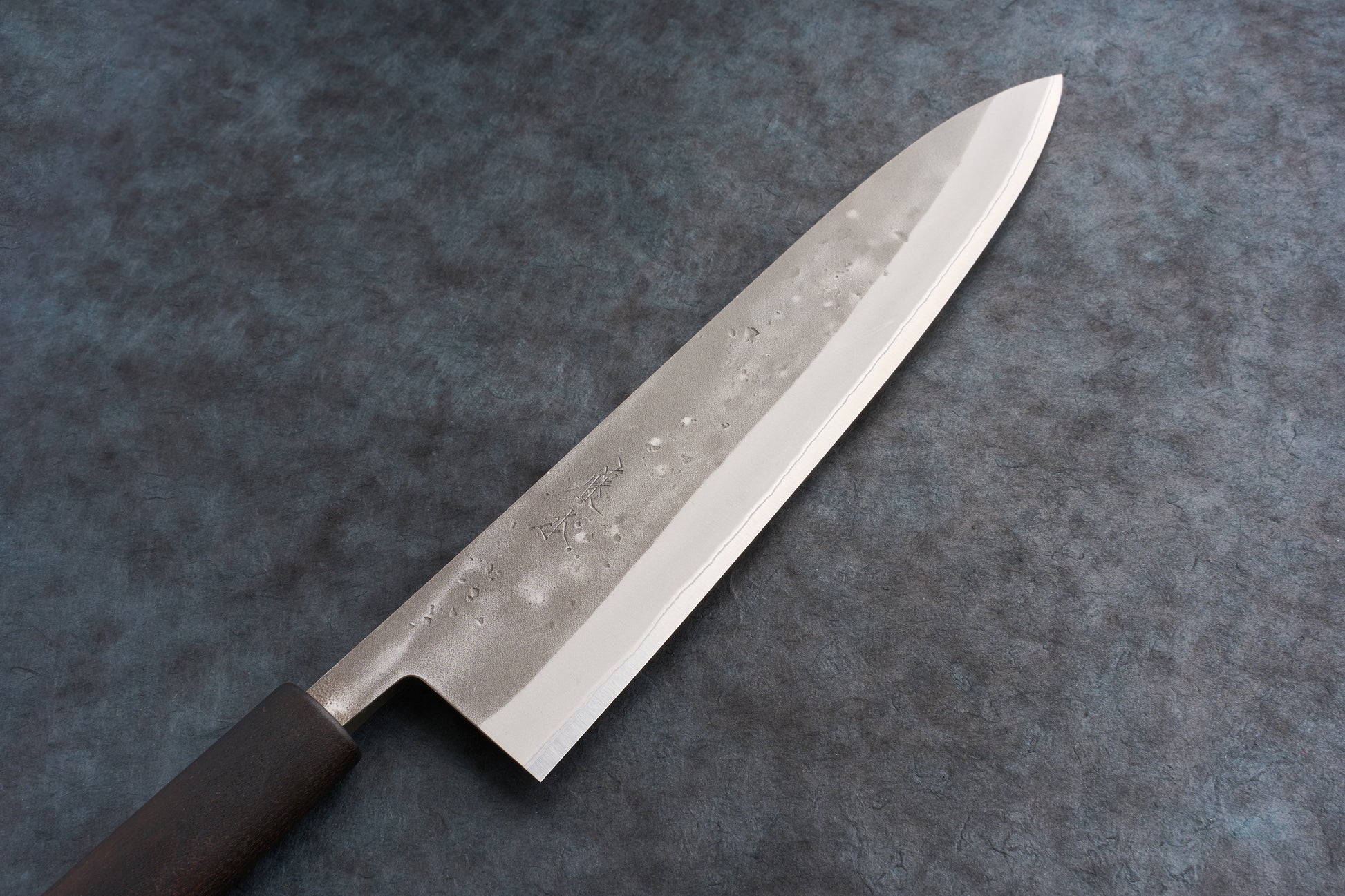 Fujimoto Nashiji Gyuto 240mm knife with a hammered finish, lying flat. The blade features a pronounced cutting edge and a visible dark handle, embodying the craftsmanship valued at Knifewear.