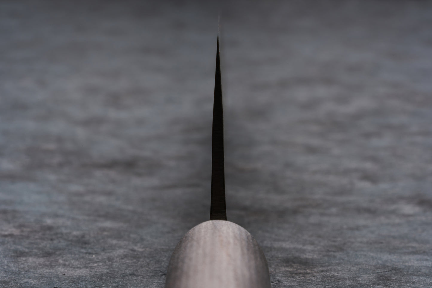 Fujimoto Nashiji Gyuto 240mm knife, showcasing its sleek metal blade and light wooden handle against a mottled grey background, exemplifies the craftsmanship and unique blend of carbon and stainless steel.