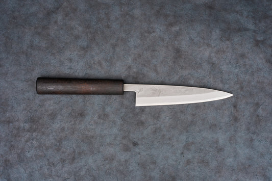 Fujimoto Nashiji Petty 135mm knife with a slender, stainless-clad carbon-steel blade and dark wooden wa-handle, lies on a gray surface, showcasing its craftsmanship ideal for precise cutting tasks.