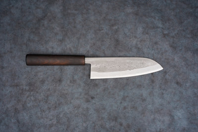 Fujimoto Nashiji Santoku 165mm, featuring a hammered blade texture with a sharp edge, rests on a gray surface. Its dark wooden wa handle emphasizes balance, embodying expert Japanese craftsmanship.