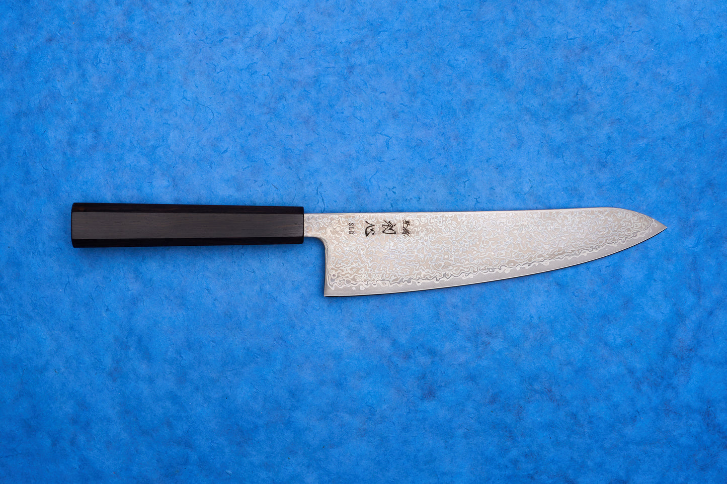 Hatsukokoro SLD Hikari Kurozome Damascus Gyuto 210mm knife with a Damascus-pattern blade and dark wooden handle on a blue background.