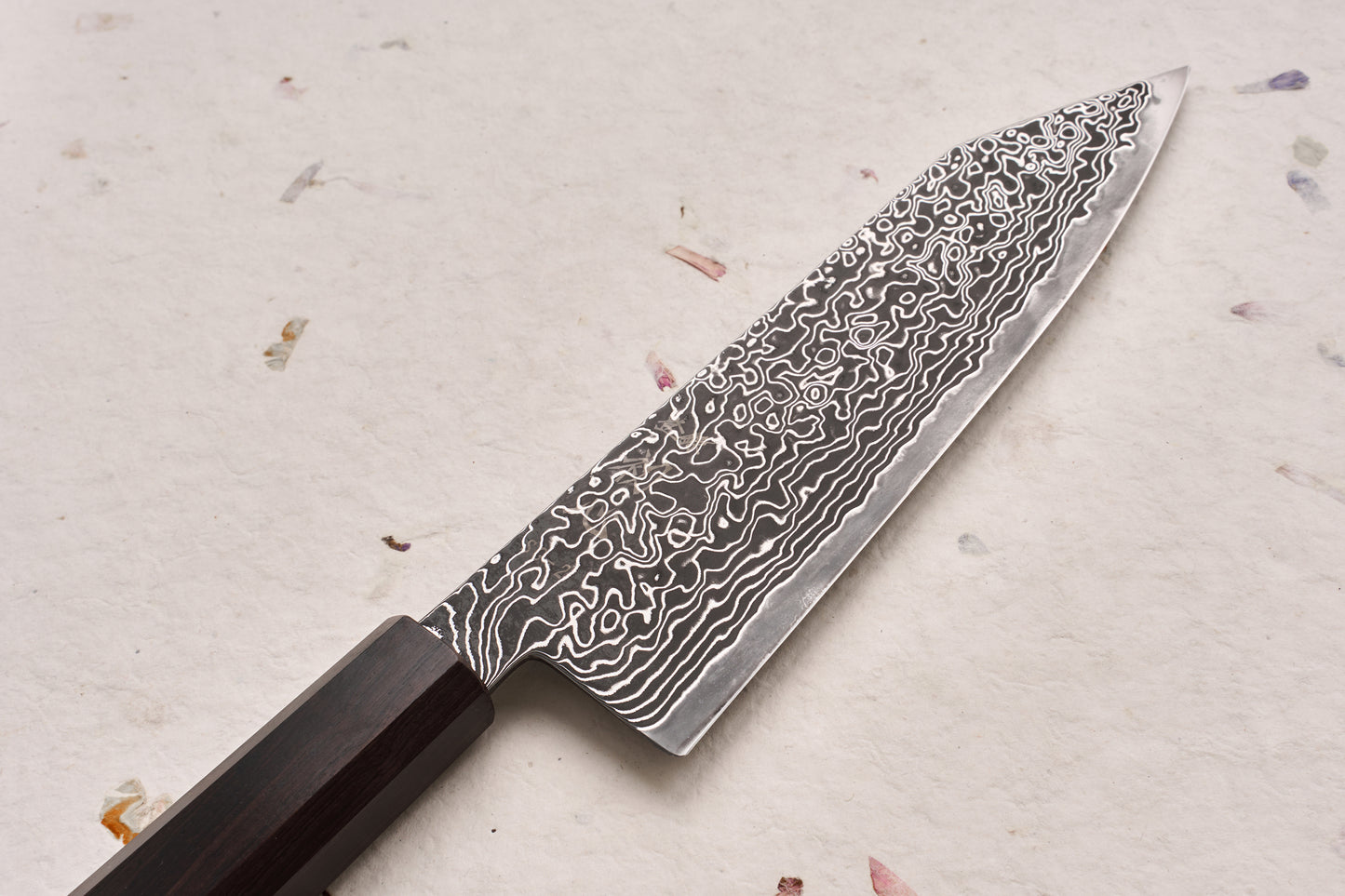 Damascus-steel Hatsukokoro Ryuhyo SG2 Kurozome Bunka knife, showcasing a swirling blade pattern and dark wood handle, lies on a textured, handmade-paper surface with embedded plant fibers.