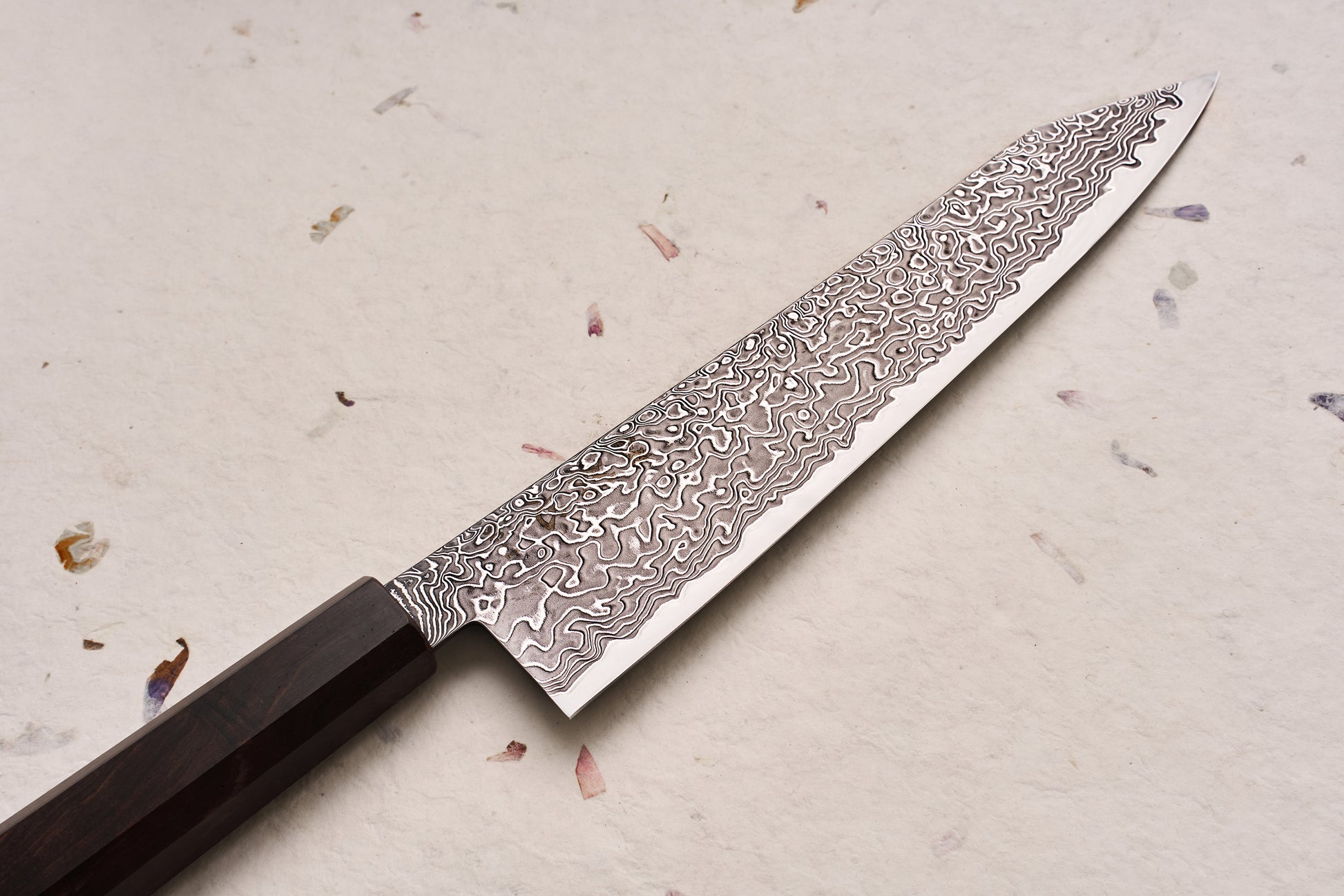 Hatsukokoro Ryuhyo SG2 Kurozome Damascus Kiritsuke Gyuto 210mm, featuring an elegantly patterned Damascus blade and a dark octagonal handle, resting on a textured surface with scattered dried flower petals.