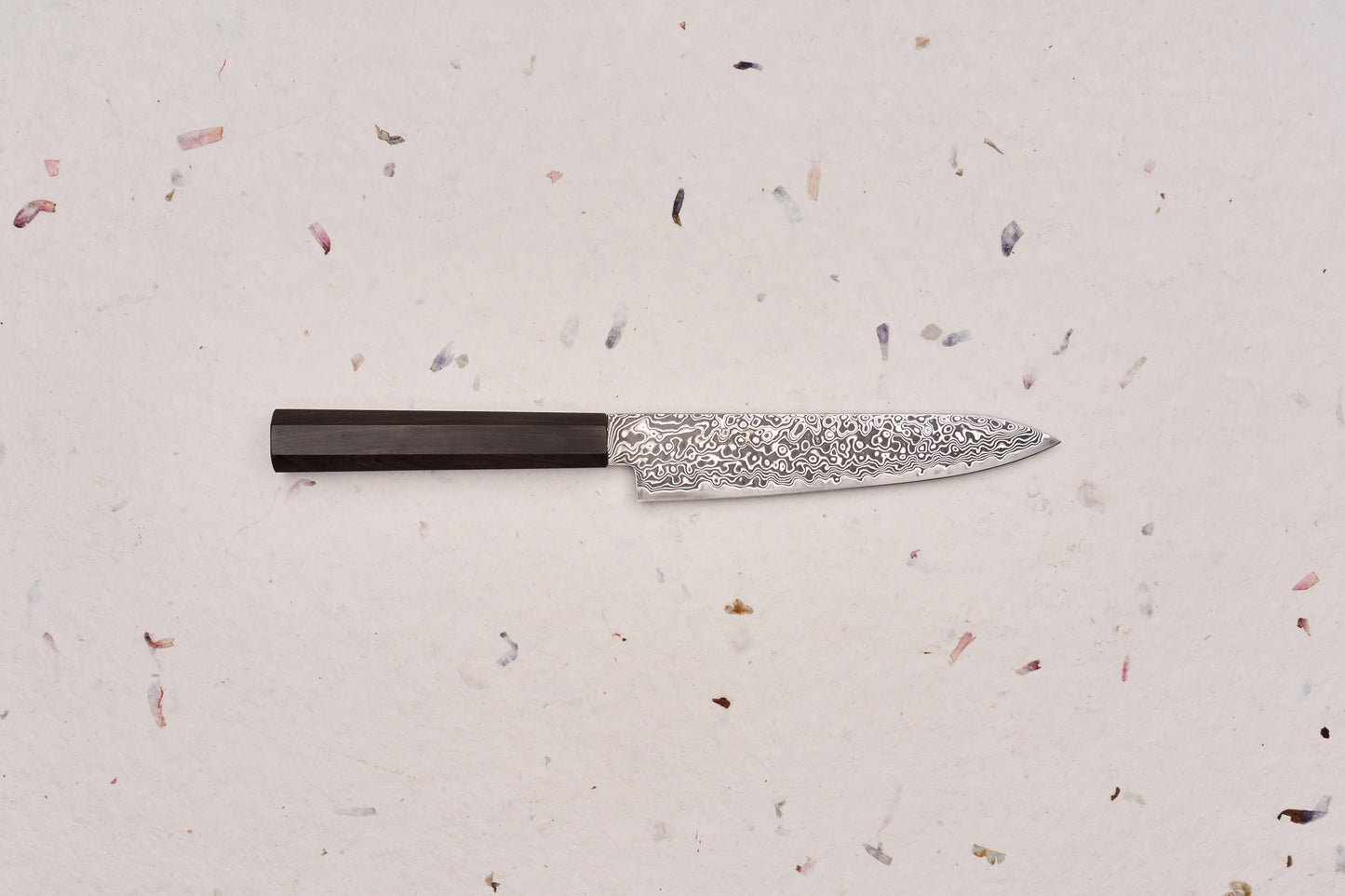Hatsukokoro Ryuhyo SG2 Kurozome Damascus Petty 150mm knife, featuring a patterned Damascus steel blade and an octagonal dark wooden handle, ideal for precise cutting tasks on a board.