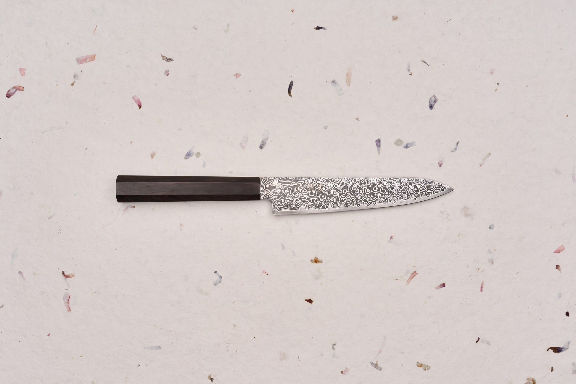 Hatsukokoro Ryuhyo SG2 Kurozome Damascus Petty 150mm knife, featuring a patterned Damascus steel blade and an octagonal dark wooden handle, ideal for precise cutting tasks on a board.