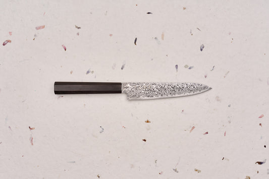 Hatsukokoro Ryuhyo SG2 Kurozome Damascus Petty 150mm knife, featuring a patterned Damascus steel blade and an octagonal dark wooden handle, ideal for precise cutting tasks on a board.