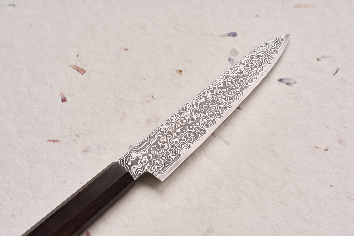 Hatsukokoro Ryuhyo SG2 Kurozome Damascus Petty 150mm knife on textured surface, showcasing its swirling Damascus blade and dark octagonal handle, embodying precision and artisanal craftsmanship.