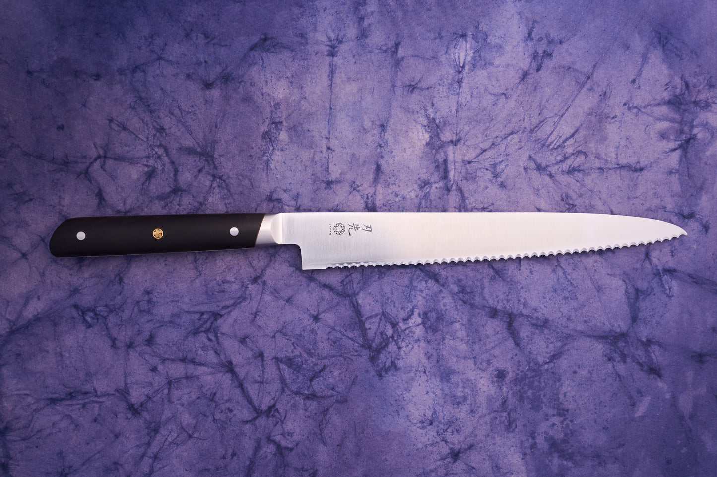 Hazaki Classic Black Bread Knife 240mm features a polished scalloped steel blade with a black handle and brass emblem, showcasing Japanese craftsmanship on a purple marbled fabric backdrop.