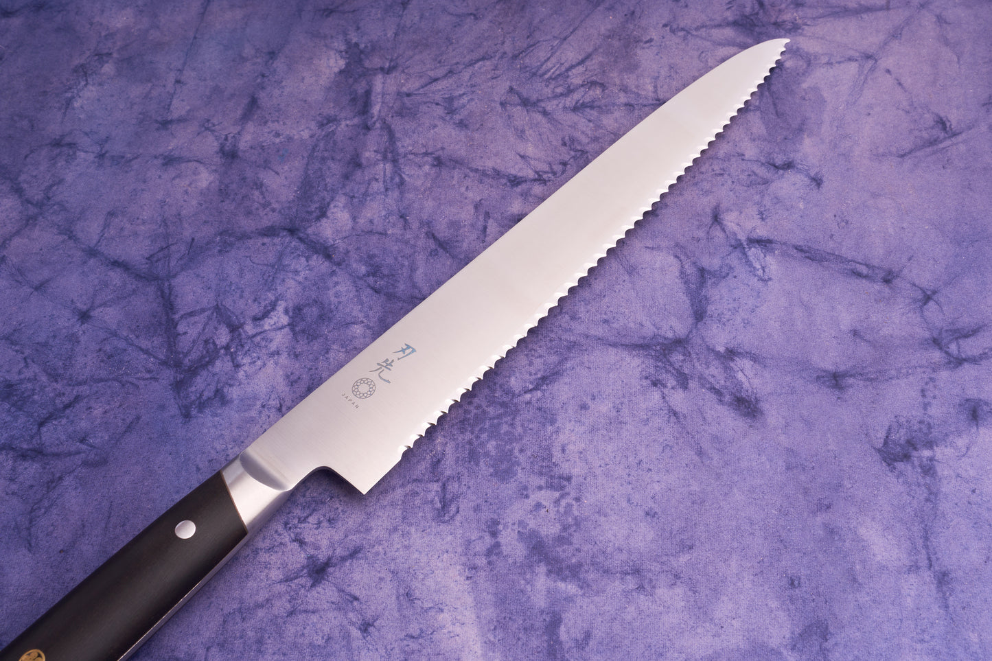 Hazaki Classic Black Bread Knife 240mm with a serrated blade inscribed with Japanese characters, laid on a mottled purple cloth. The knife showcases Japanese craftsmanship paired with Canadian woodwork.