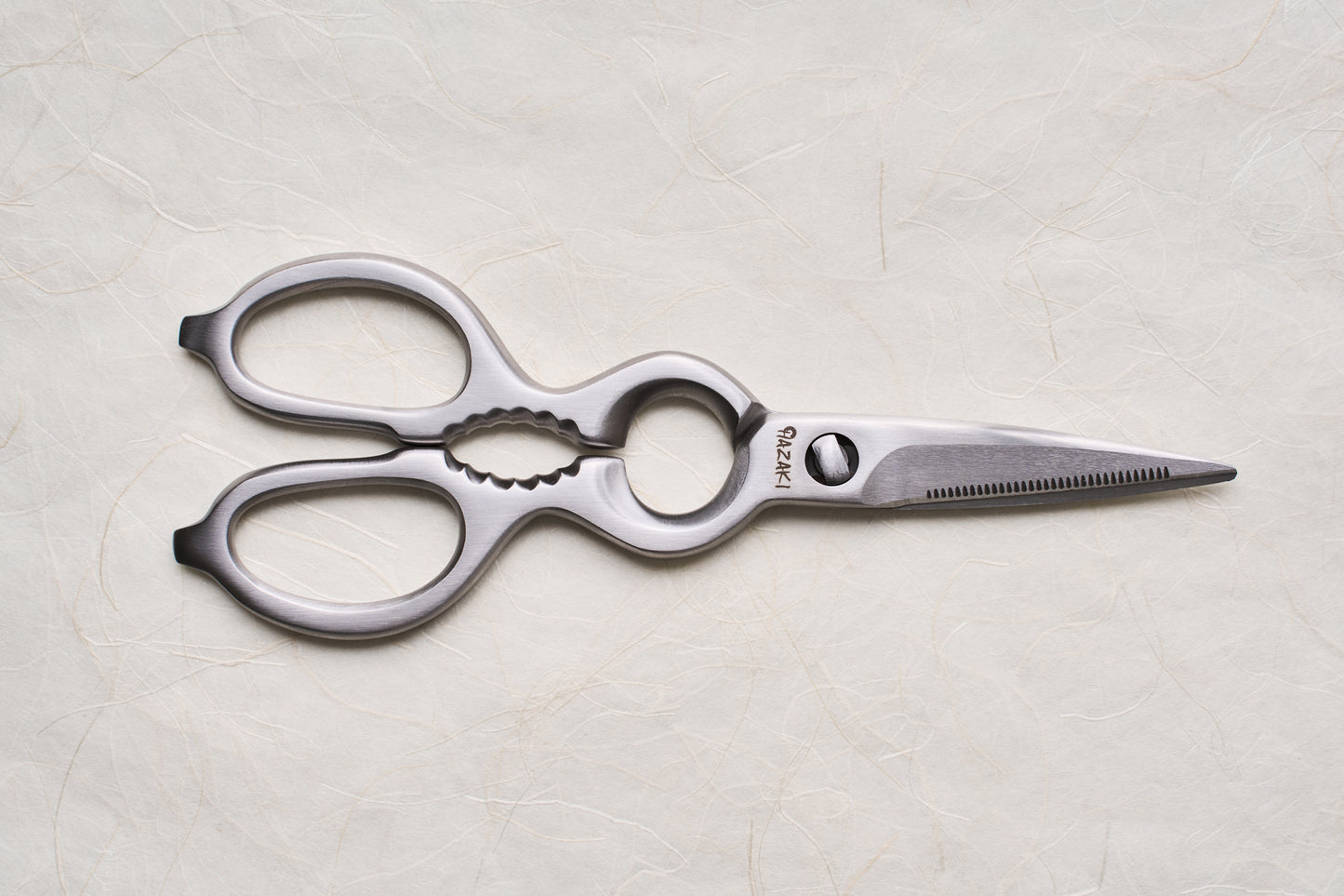 Hazaki Stainless Kitchen Shears with ergonomic handles and a serrated blade, featuring a bottle opener and nutcracker, displayed on a textured surface. Ideal for versatile kitchen tasks and easy maintenance.