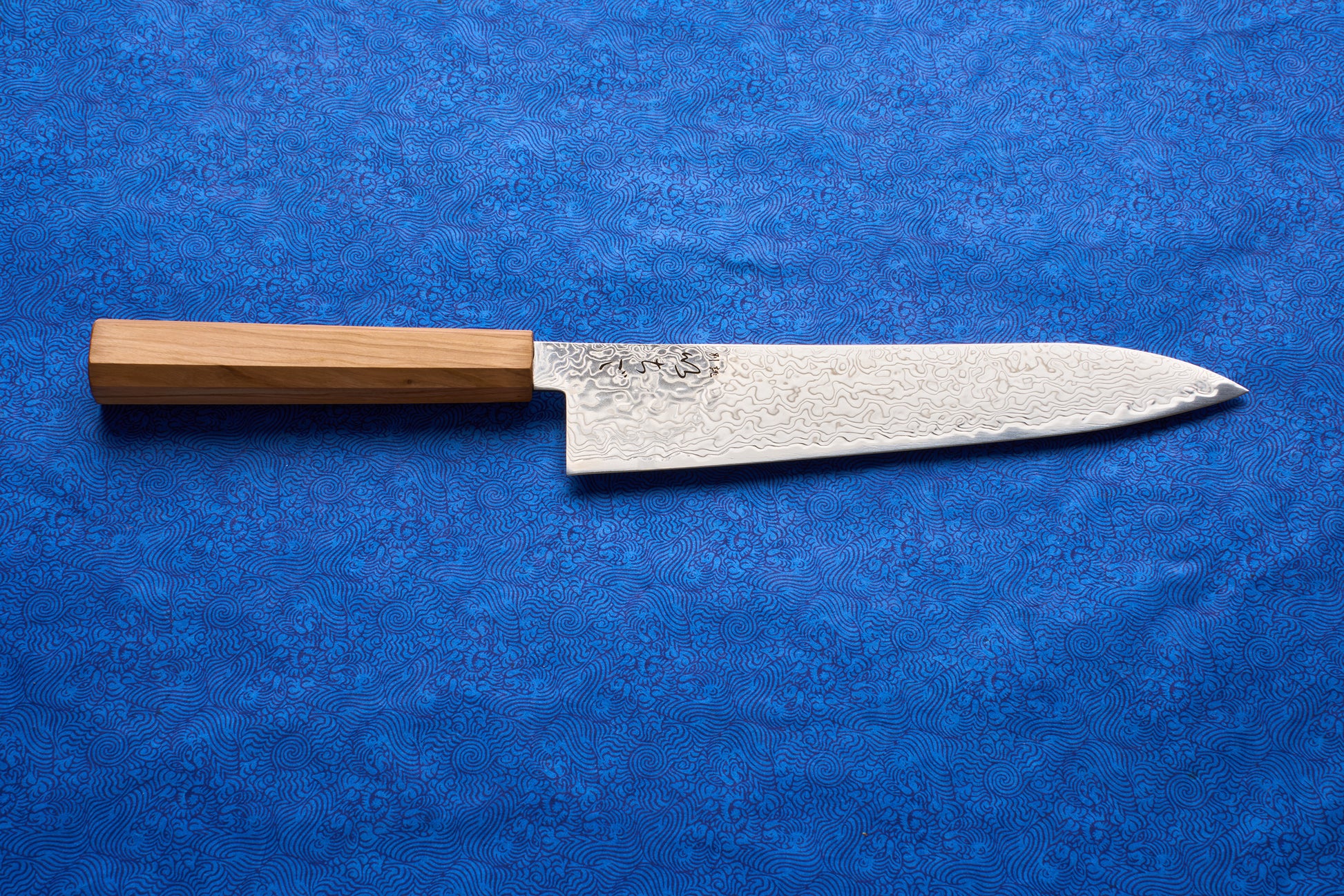 ICHIGO Haku Gyuto 210mm chef's knife on a blue cloth, showcasing a Damascus-style wave-patterned blade and a lightweight wooden wa-handle, embodying Japanese craftsmanship and premium performance.