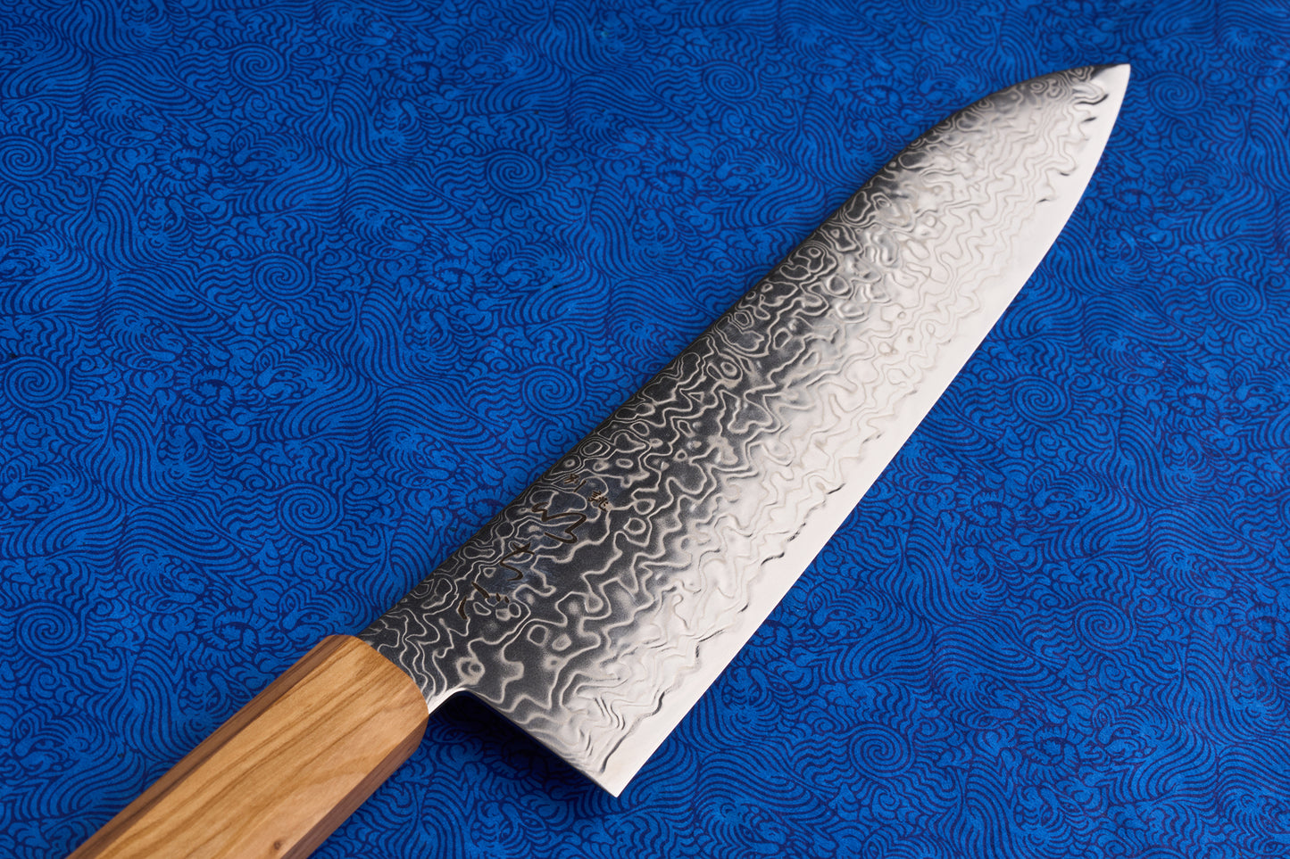 ICHIGO Haku Gyuto 210mm features a striking Damascus steel blade and light wooden handle, set against a swirling blue fabric, embodying Knifewear's dedication to exquisite, high-performance Japanese kitchen knives.