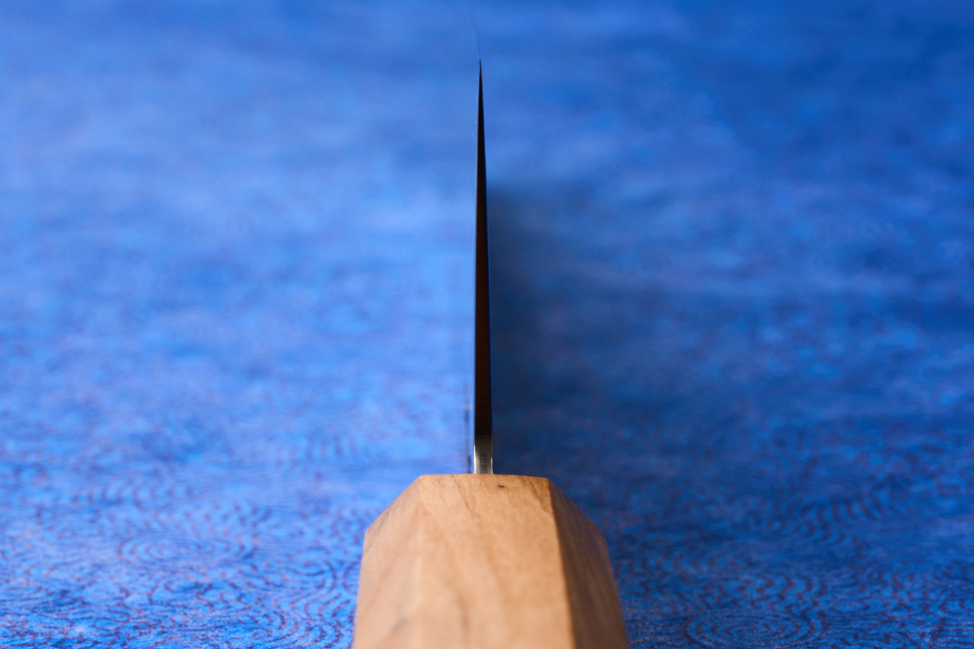 ICHIGO Haku Gyuto 210mm knife features a sleek, super-thin steel blade emerging from a faceted wooden handle, resting on a subtly patterned blue surface, epitomizing high-performance Japanese craftsmanship.
