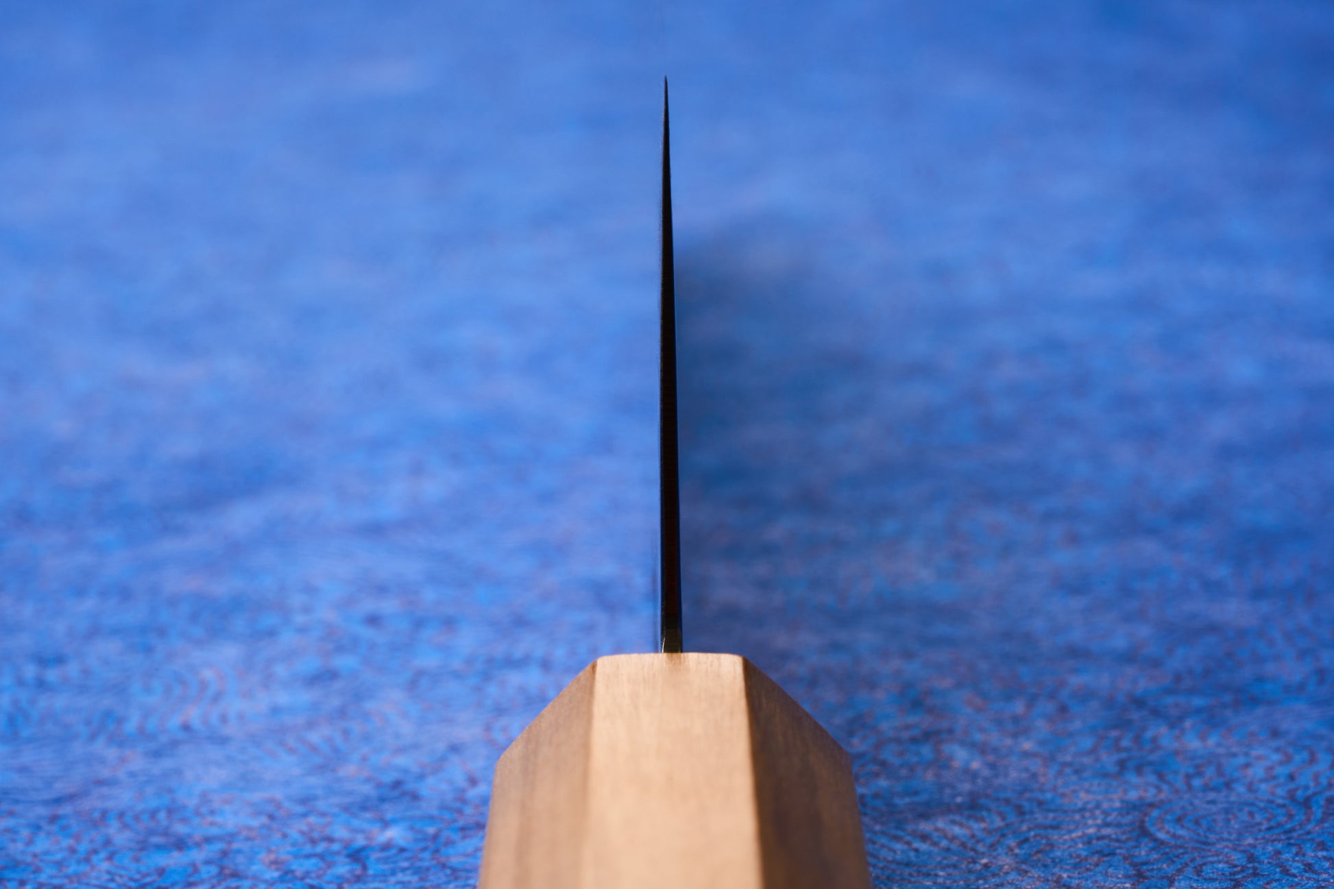 ICHIGO Haku Gyuto 240mm stands vertically, showcasing its thin metal blade and light wooden handle, set against a softly blurred blue background, embodying precision and elegance for culinary enthusiasts.