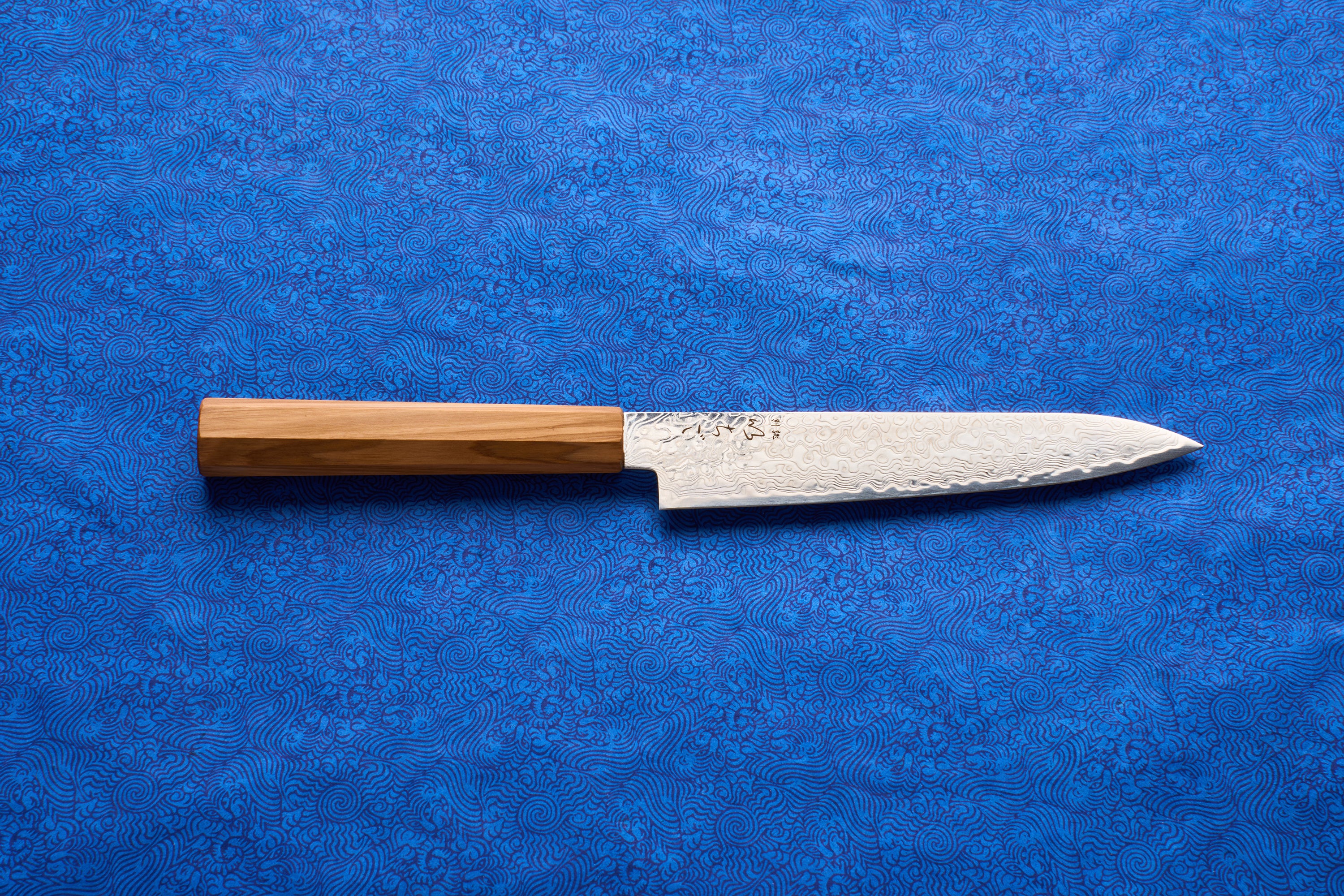 ICHIGO Haku Petty 150mm knife with a Damascus-pattern blade and a straight wooden handle, displayed on a blue patterned surface, ideal for precise cutting tasks like slicing shallots and herbs.