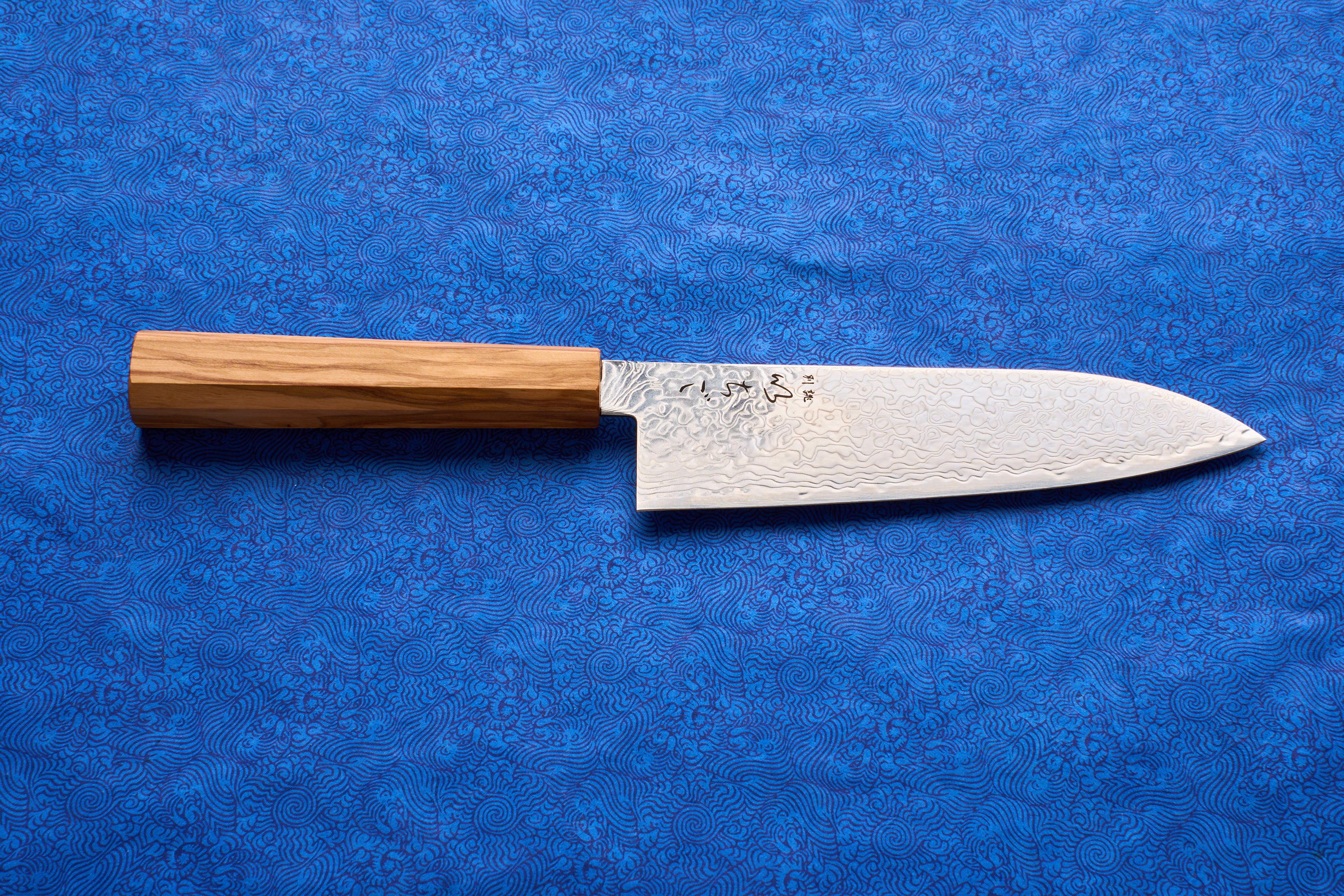 ICHIGO Haku Santoku 180mm knife on a blue cloth, featuring a wavy Damascus steel blade with tsuchime texture and Japanese characters, paired with a light wooden handle, ideal for versatile kitchen tasks.