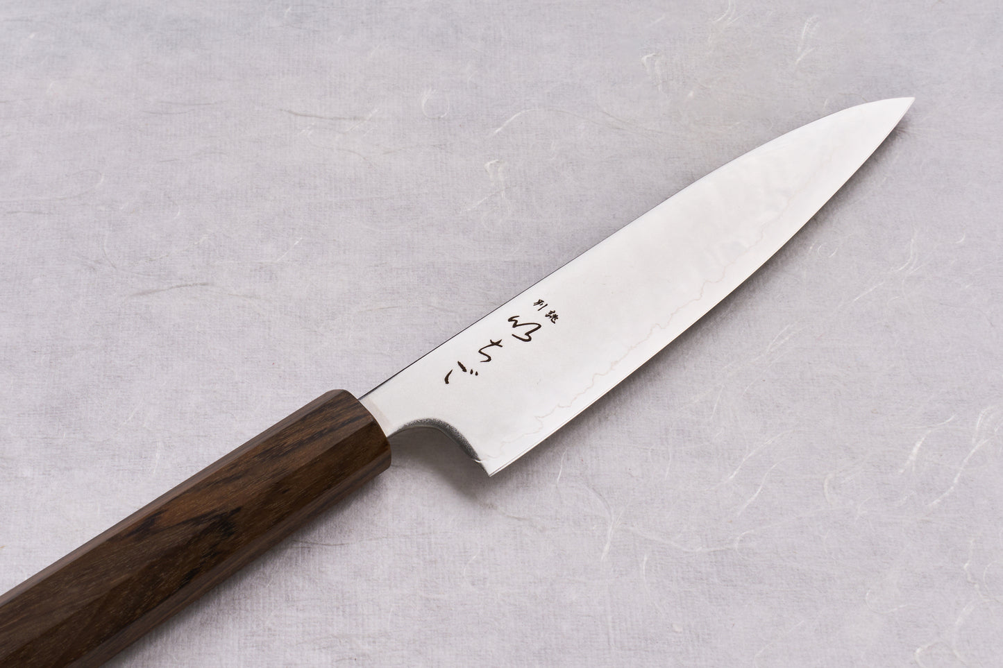 ICHIGO SLD Migaki Petty 130mm knife with a slim stainless-steel blade featuring Japanese characters and a dark wood octagonal handle, displayed on a pale, textured surface.