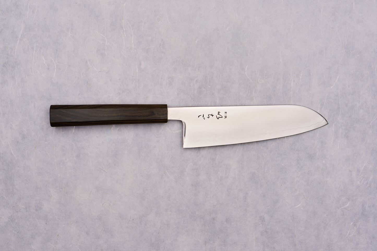 ICHIGO SLD Migaki Santoku 170mm lies flat, showcasing its polished steel blade with Japanese characters and dark wooden handle, embodying timeless craftsmanship and precision, ideal for versatile kitchen tasks.