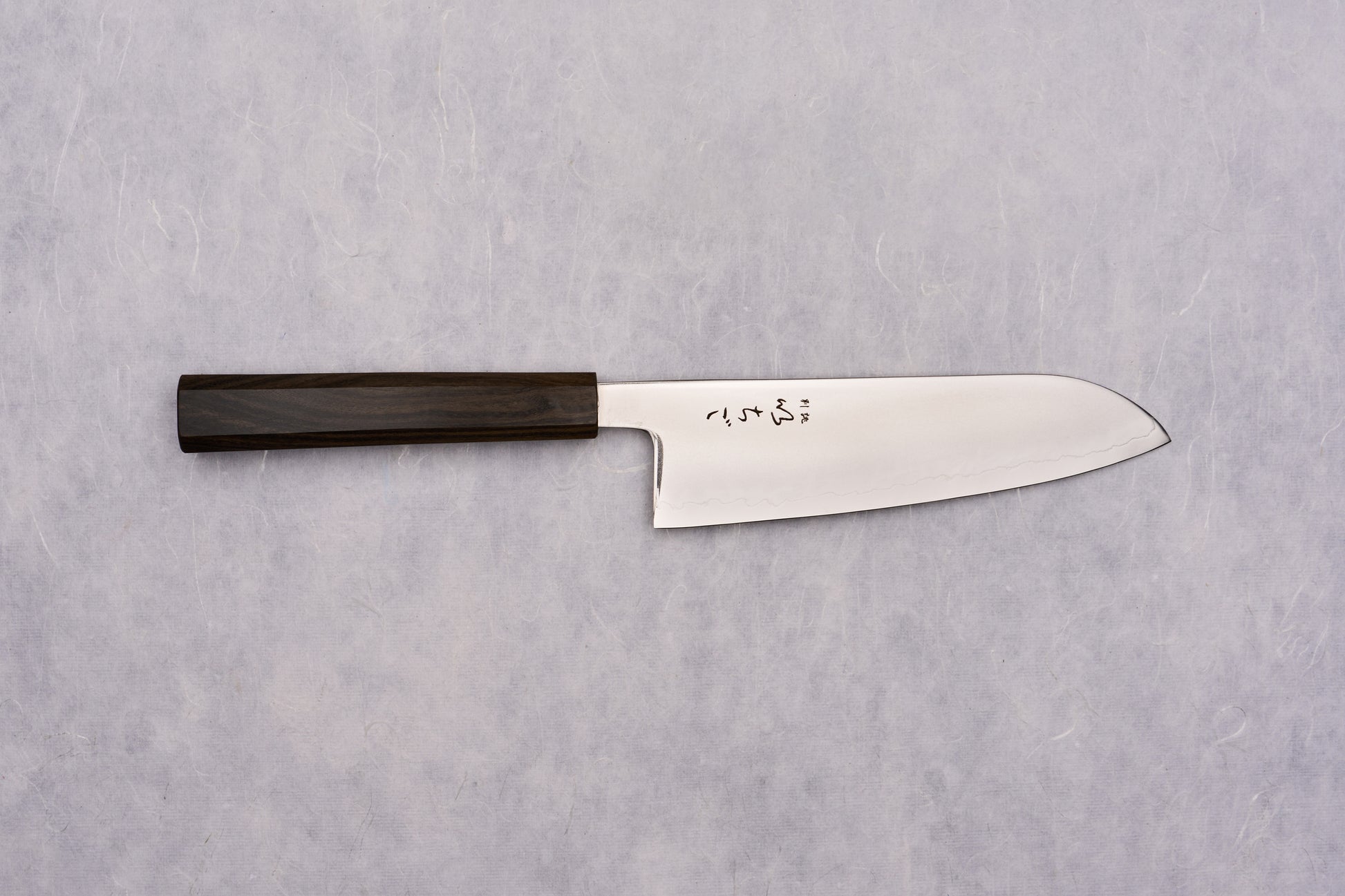 ICHIGO SLD Migaki Santoku 170mm lies flat, showcasing its polished steel blade with Japanese characters and dark wooden handle, embodying timeless craftsmanship and precision, ideal for versatile kitchen tasks.