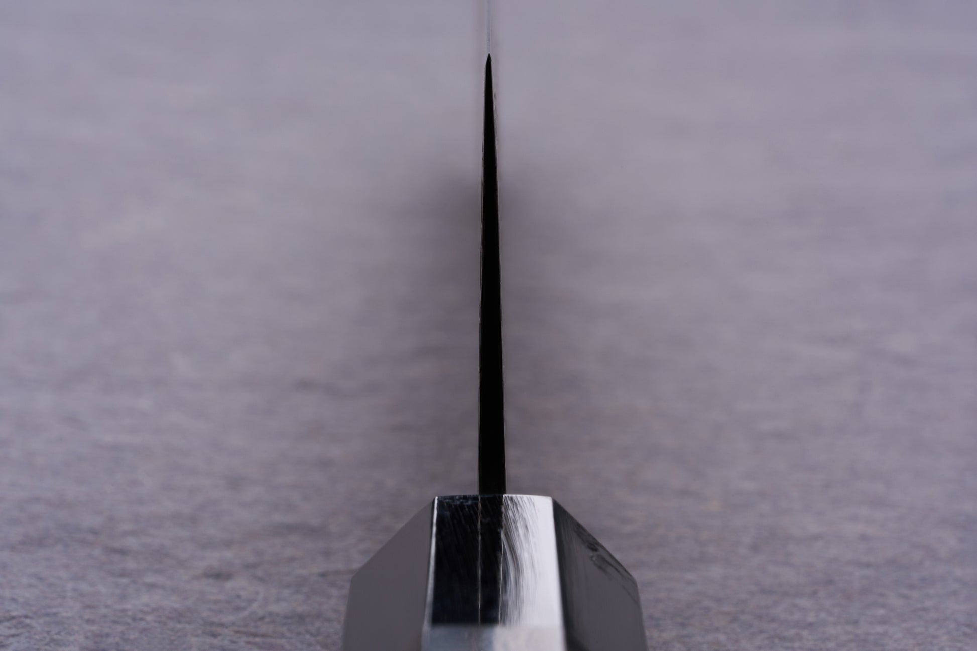 Close-up of Kanjo SG2 Nickel Damascus Kiritsuke Gyuto 240mm knife blade edge, showcasing its precision craftsmanship and sleek, polished bolster against a softly blurred tabletop backdrop.