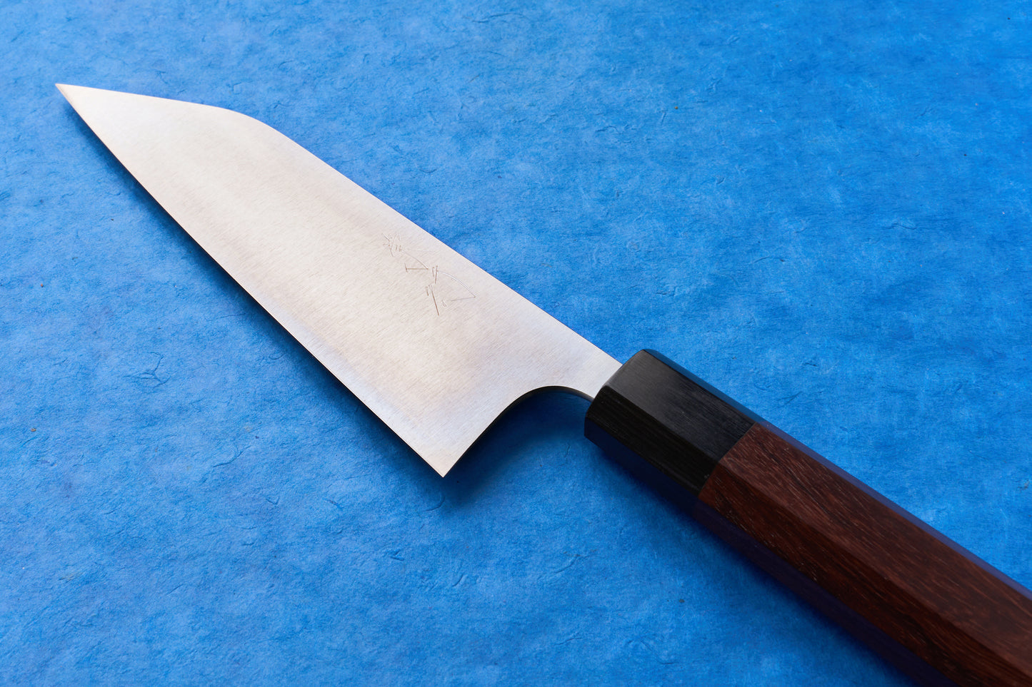 Shibata Koutetsu Chromax Migaki Santoku 165mm features a sleek stainless steel blade and octagonal wooden handle, ideal for slicing, dicing, and mincing, epitomizing Japanese craftsmanship on a blue surface.