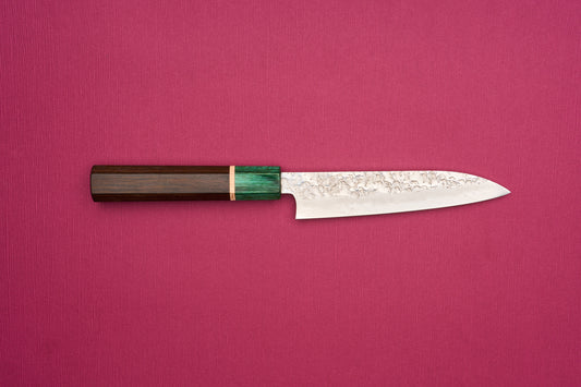 Makoto Kurosaki SPG STRIX Samidorizuki Petty 135mm knife with a hammered blade and octagonal dark wood handle, displayed on a solid magenta background, showcasing its handcrafted Japanese design.