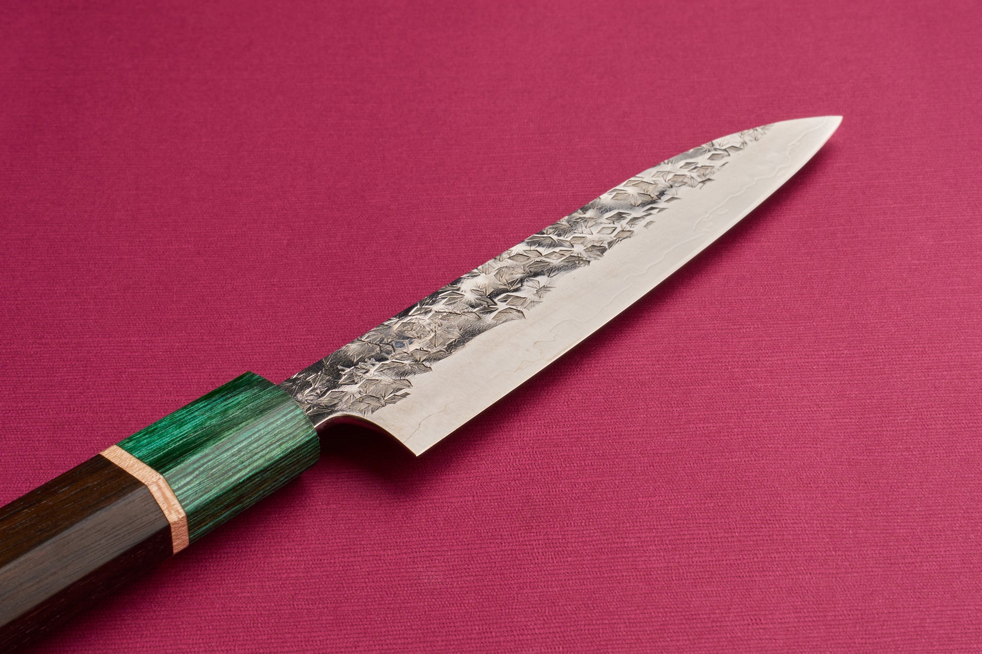 Makoto Kurosaki SPG STRIX Samidorizuki Petty 135mm knife on a textured surface, featuring a Damascus-style blade with a hammered pattern and an octagonal dark wood handle with a green ferrule.