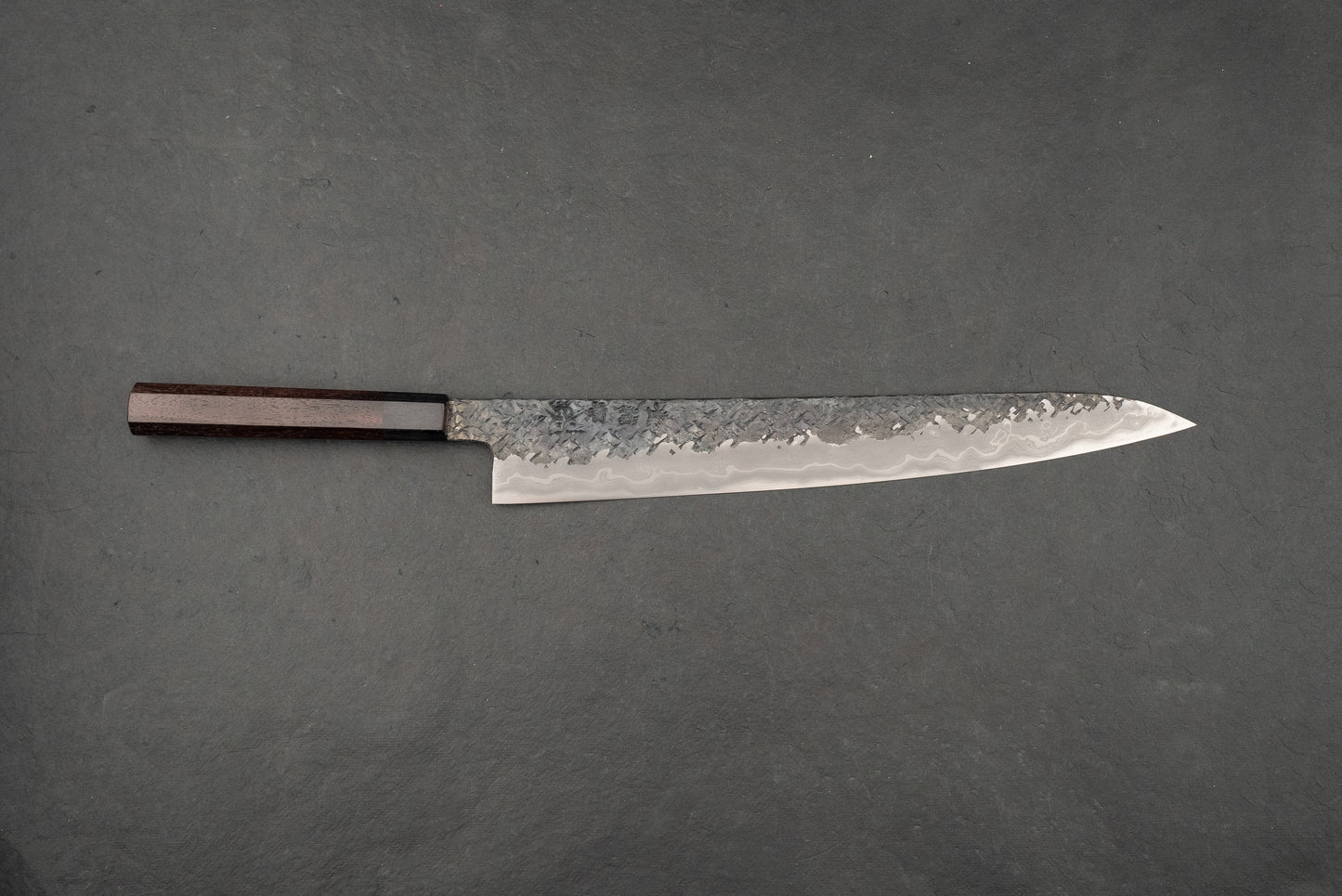Kisuke Manaka ENN Sujihiki 300mm knife features a hammered Damascus-patterned blade and an octagonal wooden handle, showcasing masterful craftsmanship on a dark slate surface, embodying Japanese blacksmithing excellence.