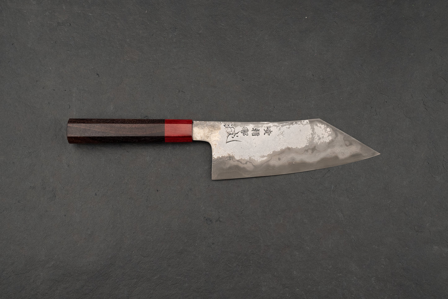 Kisuke Manaka KOKUENN Hakata 180mm knife, featuring a Damascus-patterned steel blade with a hamon-like line and stamped Japanese characters, paired with a dark octagonal handle and bright red ferrule.